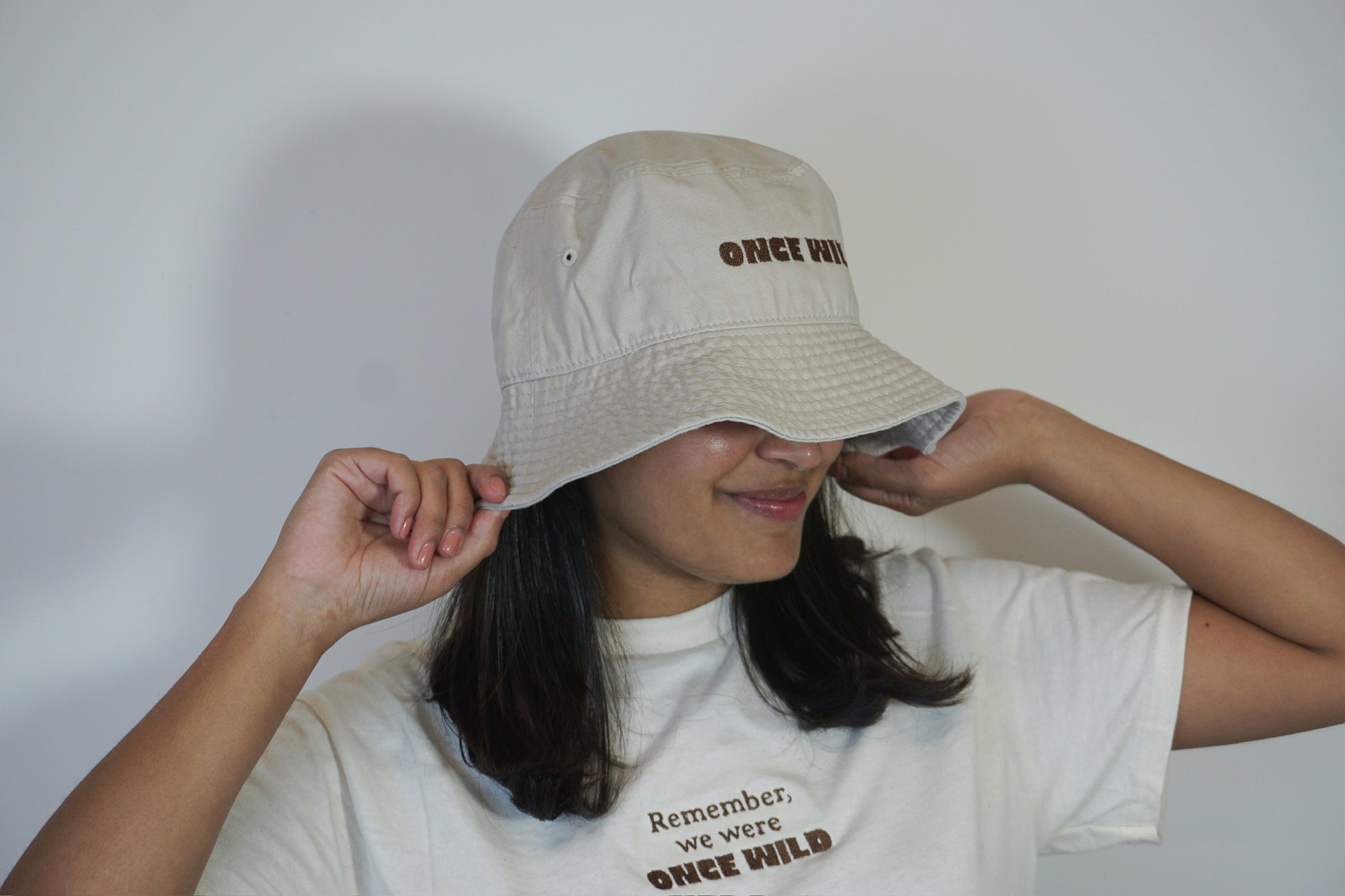 Person wearing a beige bucket hat and white t-shirt with text against a plain background