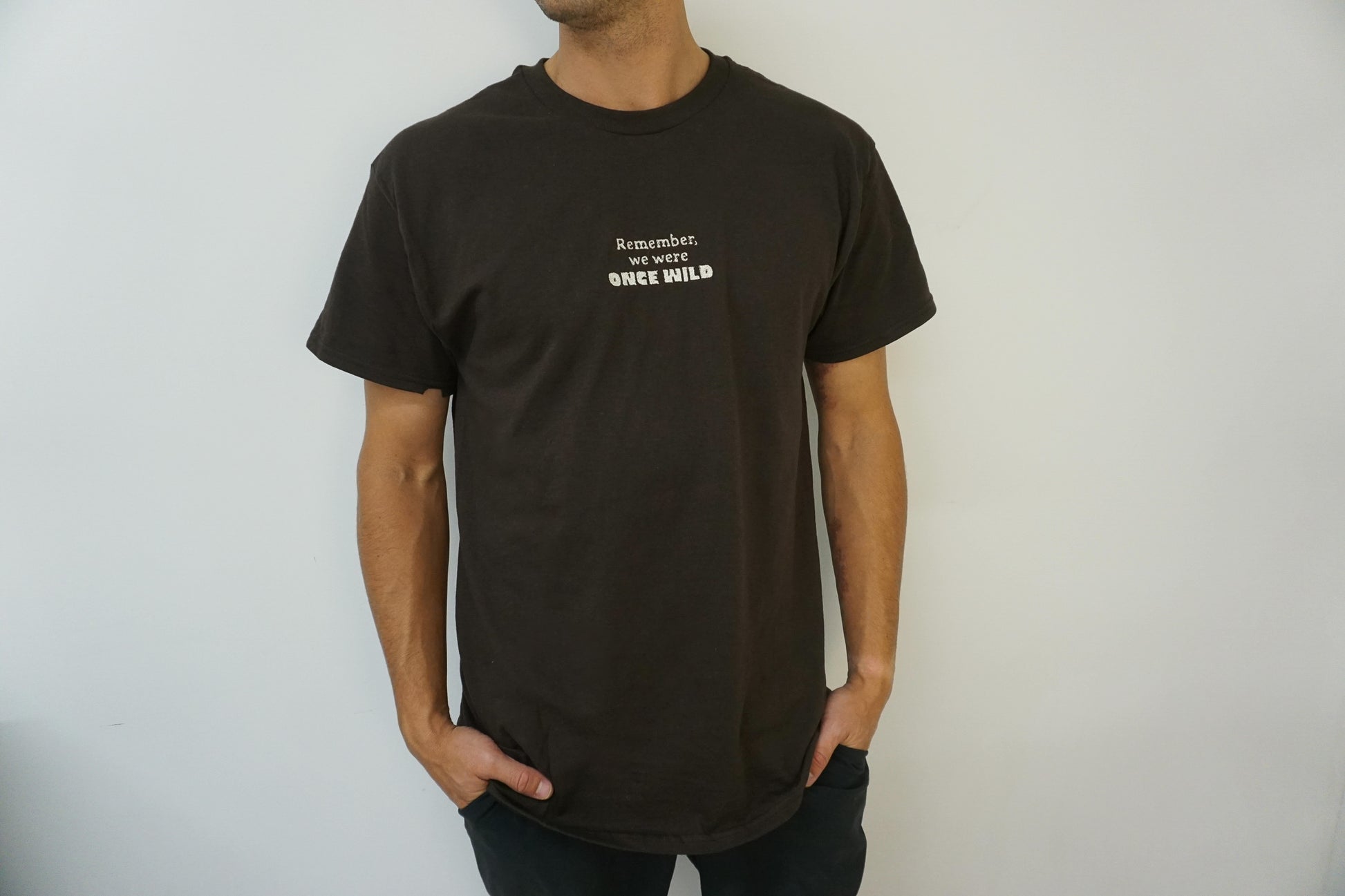 Man wearing a brown t-shirt with text on a plain background that says "Remember, we were Once Wild" 